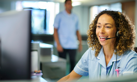 A medical office assistant sits at a desk with a headset, talking to a patient on the phone. She is looking at a computer screen and smiling.